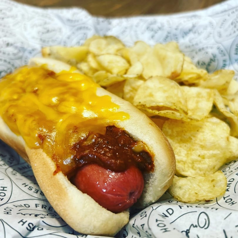 A chili cheese hot dog with a side of potato chips, served in a paper-lined basket.