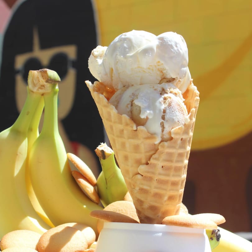 Two scoops of banana pudding ice cream in a waffle cone, with bananas and vanilla wafers.