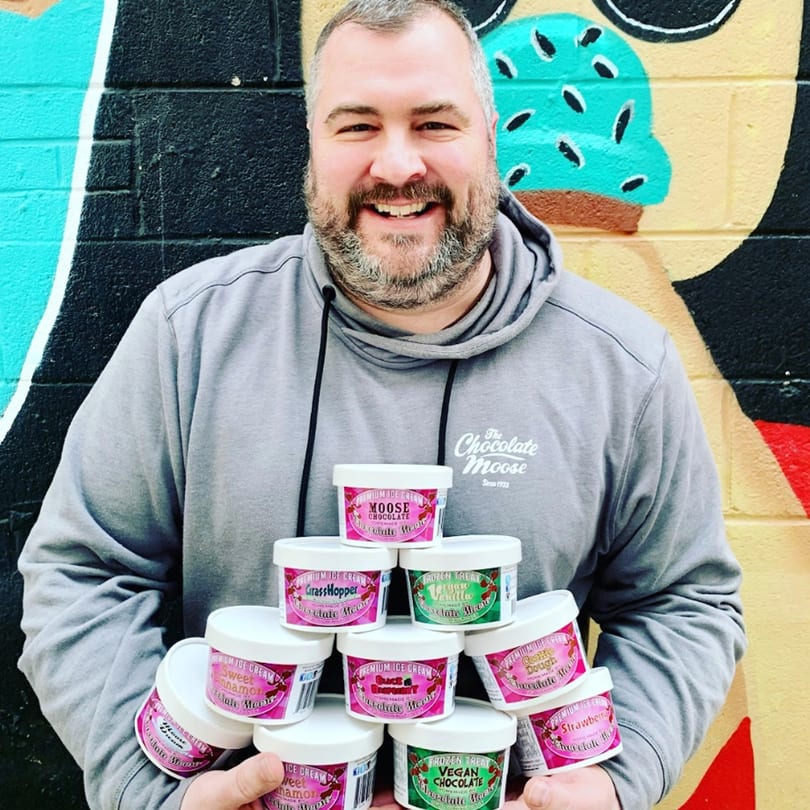 A smiling man in a grey hoodie holds a stack of Chocolate Moose ice cream tubs against a colorful mural.