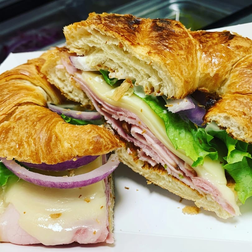 A croissant sandwich with ham, melted cheese, lettuce, and red onion, cut in half.